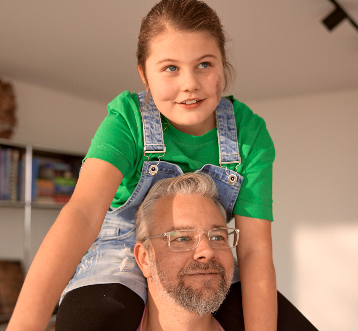 Tochter sitzt auf den Schultern ihres Vaters. Sie hat ein grünes T-shirt an und eine Latzhose